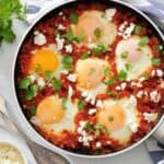 A pan of shakshuka with poached eggs, tomato sauce, crumbled cheese, and cilantro, placed on a striped towel with fresh herbs, tomatoes, and a small bowl of cheese nearby.