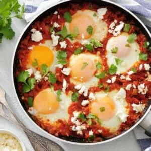 A pan of shakshuka with poached eggs, tomato sauce, crumbled cheese, and cilantro, placed on a striped towel with fresh herbs, tomatoes, and a small bowl of cheese nearby.