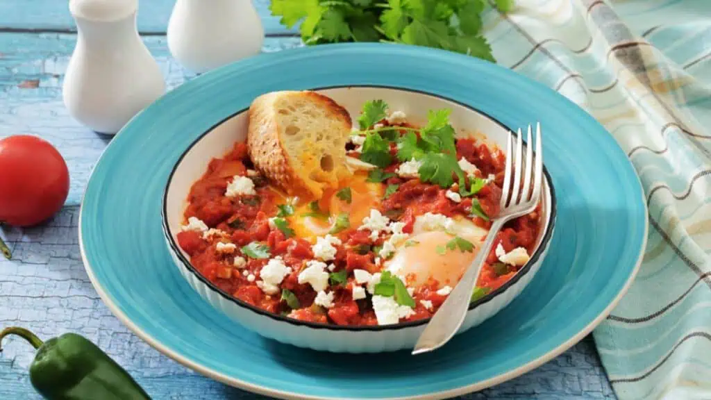 A plate of shakshuka with poached eggs, tomato sauce, feta cheese, cilantro, and a slice of bread, served with a fork on a blue plate.