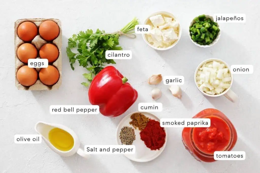 A selection of labeled ingredients on a white surface, including eggs, cilantro, feta, jalapeños, onion, garlic, red bell pepper, cumin, smoked paprika, olive oil, salt and pepper, and tomatoes.