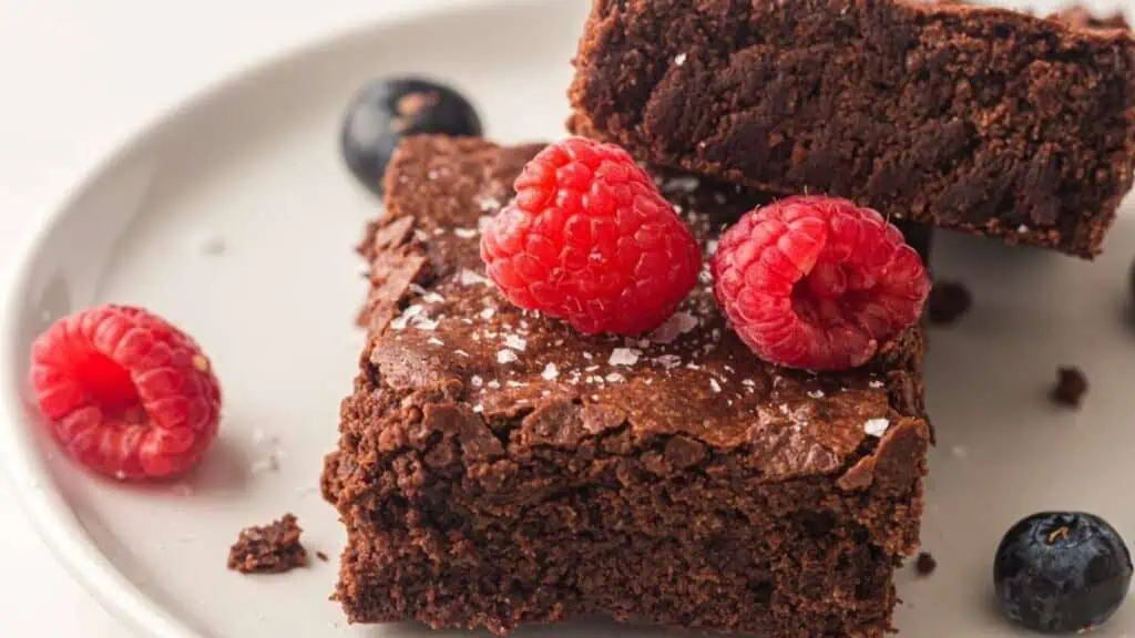 Two chocolate brownies on a white plate, topped with raspberries, with blueberries and extra raspberries on the side.