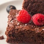 Two chocolate brownies on a white plate, topped with raspberries, with blueberries and extra raspberries on the side.