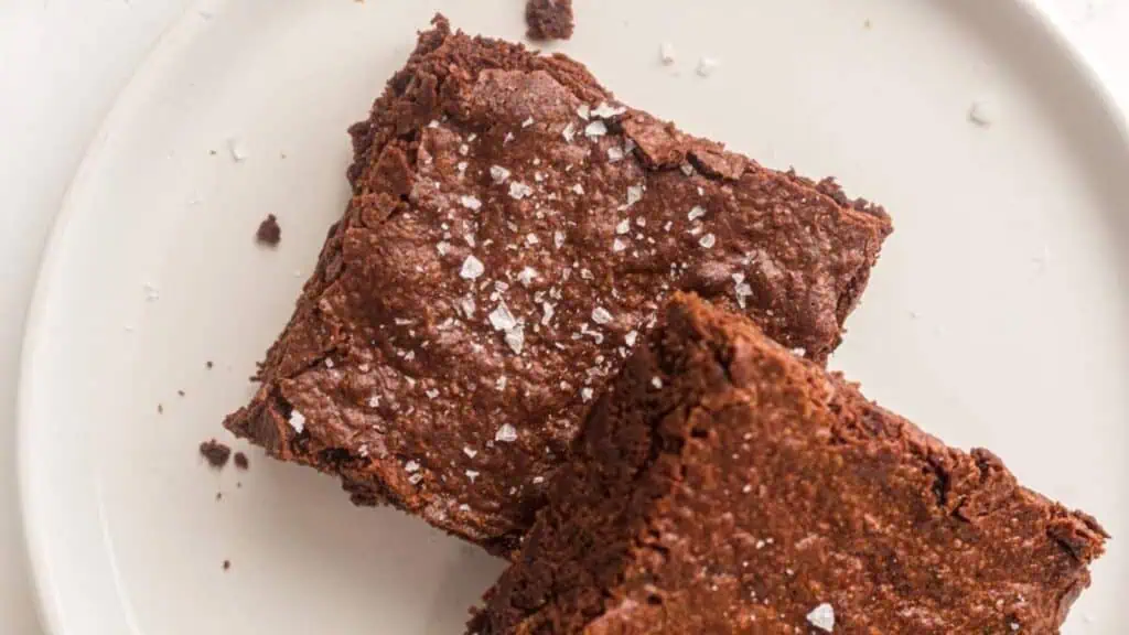 Two chocolate brownies topped with flaky sea salt on a white plate.