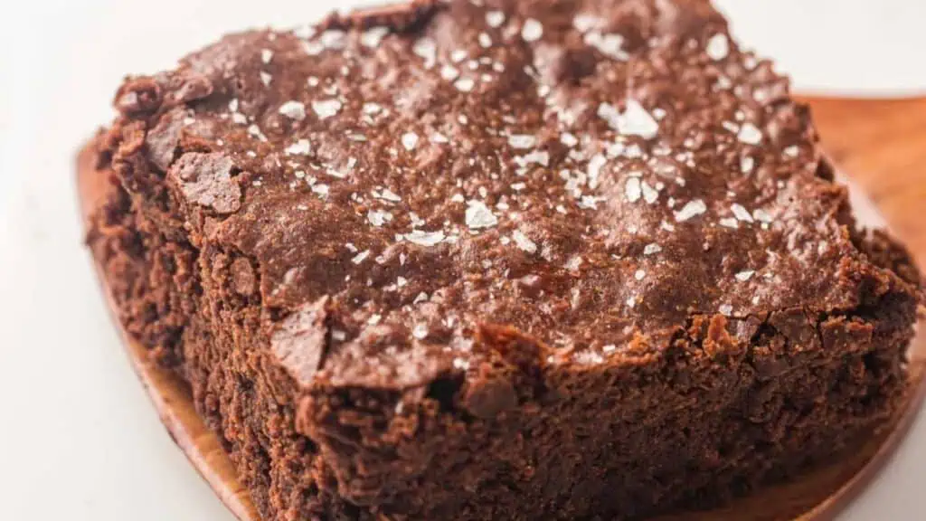 A close-up of a thick, chocolate brownie with a crinkled top and sea salt flakes sprinkled on it, resting on a wooden surface.