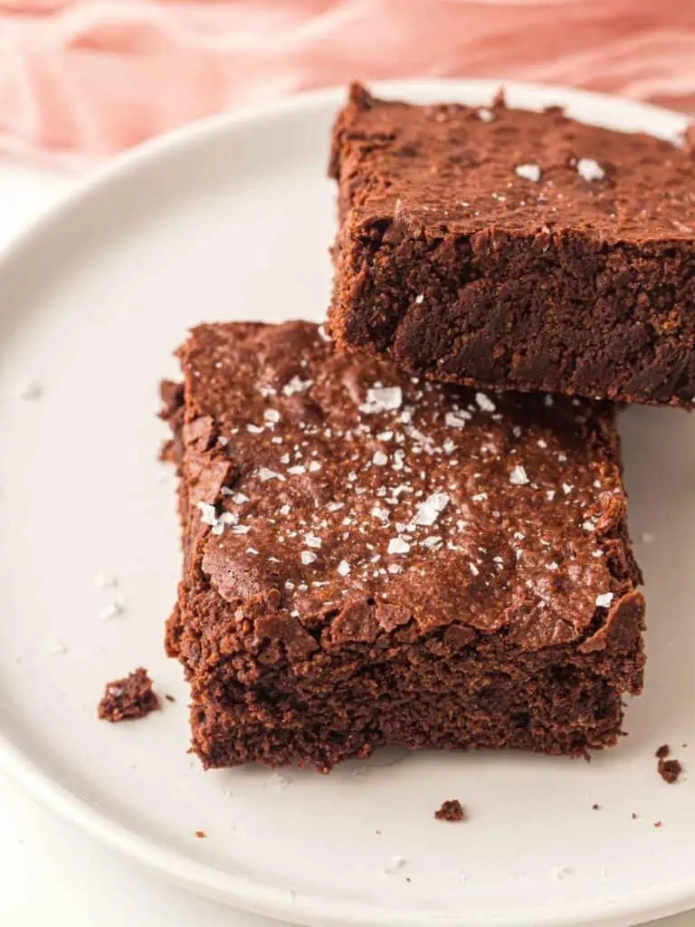 Two chocolate brownies sprinkled with flaky sea salt sit on a white plate.