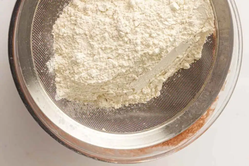 Flour being sifted through a metal mesh strainer over a bowl, with some flour already sifted below.