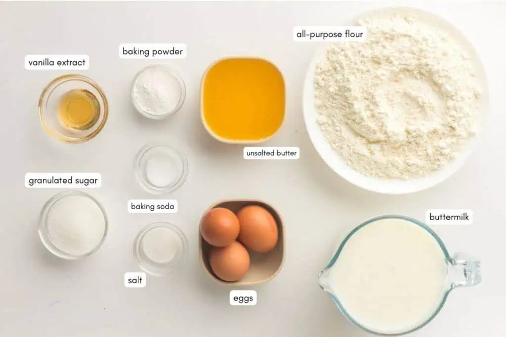 Nine baking ingredients on a white surface: vanilla extract, baking powder, melted butter, flour, sugar, baking soda, salt, three eggs, and buttermilk, each labeled with text.