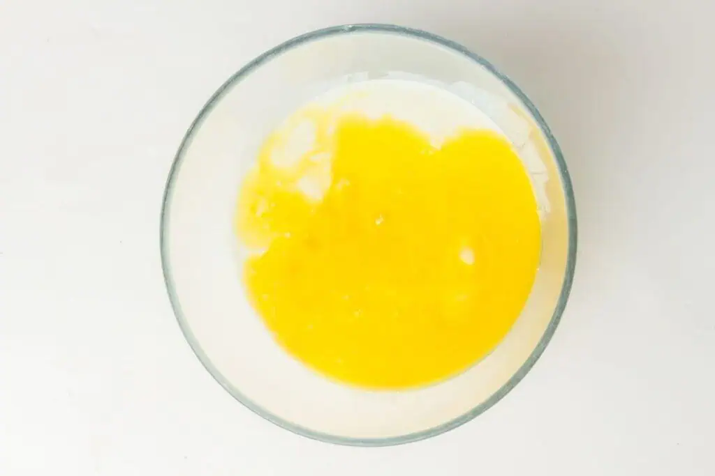 A glass bowl containing a yellow liquid mixture of eggs and milk sits on a white surface, viewed from above.