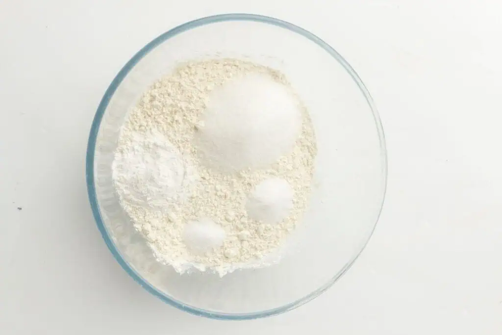 A glass bowl containing flour, sugar, and other dry baking ingredients on a white surface.