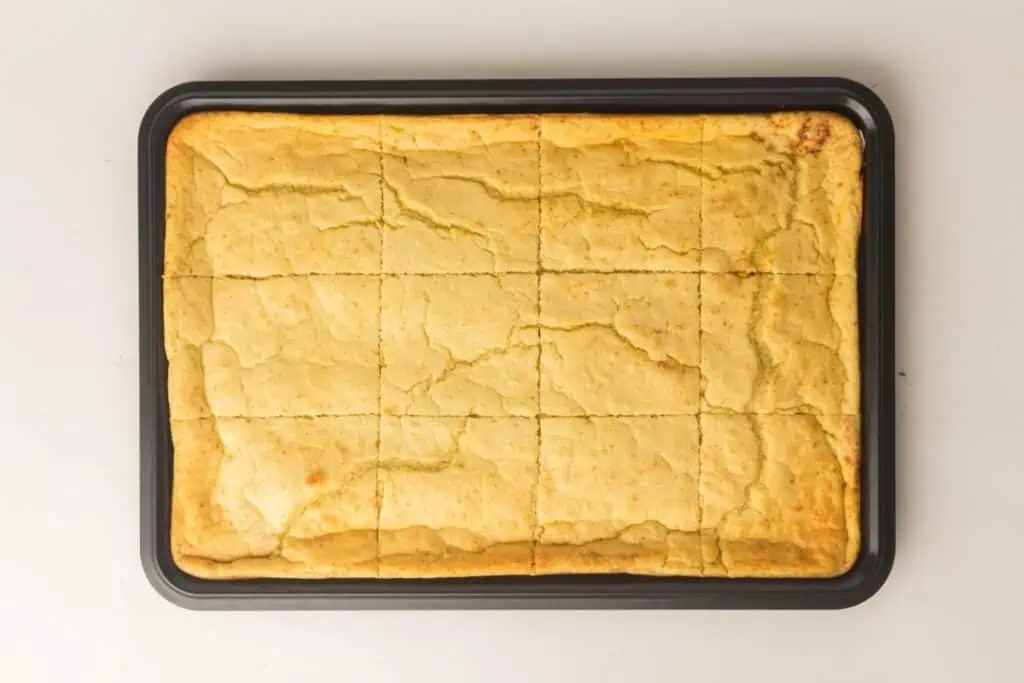 A rectangular baking tray with a large, golden-brown cornbread that is cut into even squares, resting on a white surface.
