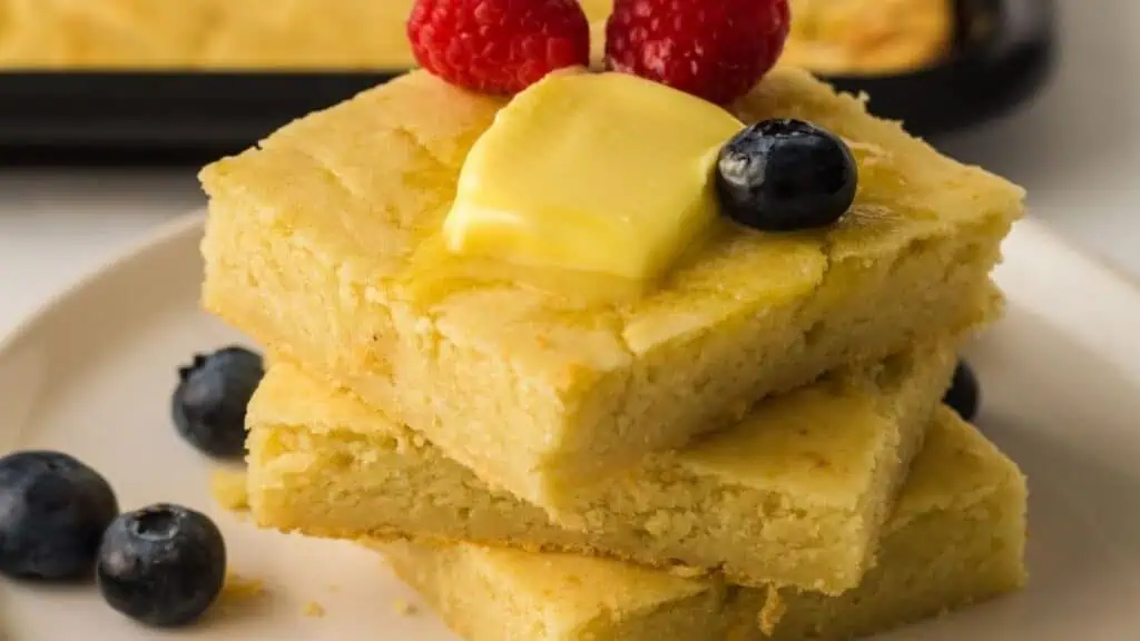 Three pieces of cornbread stacked on a plate, topped with butter, raspberries, and a blueberry, with additional blueberries on the plate.