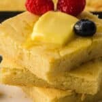 Three pieces of cornbread stacked on a plate, topped with butter, raspberries, and a blueberry, with additional blueberries on the plate.