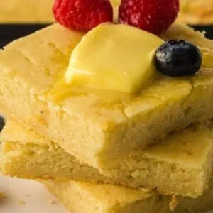 Three pieces of cornbread stacked on a plate, topped with butter, raspberries, and a blueberry, with additional blueberries on the plate.