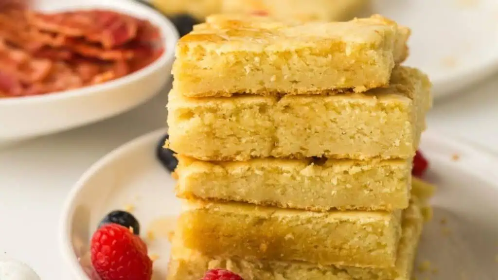 A stack of four thick, golden pancakes on a white plate, garnished with raspberries and blueberries, with a dish of bacon in the background.