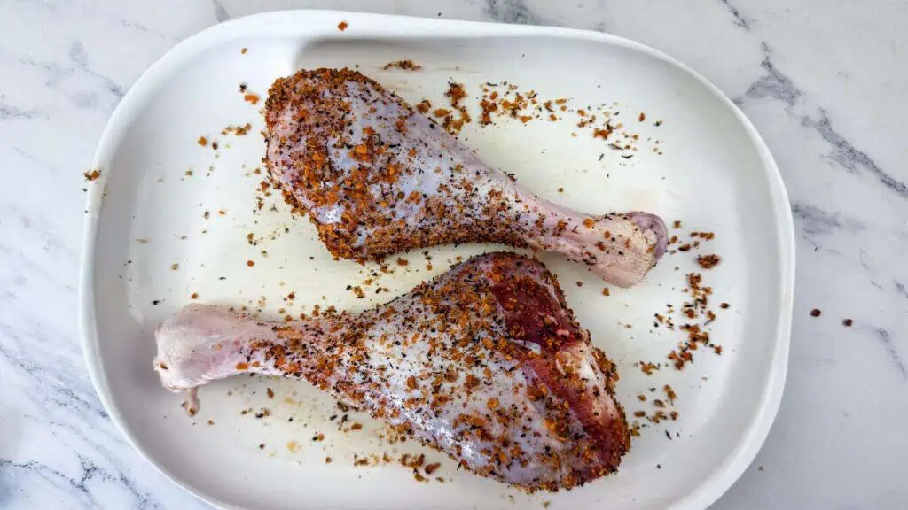 Two raw turkey legs coated with a spice rub on a white oval dish, placed on a marble surface.