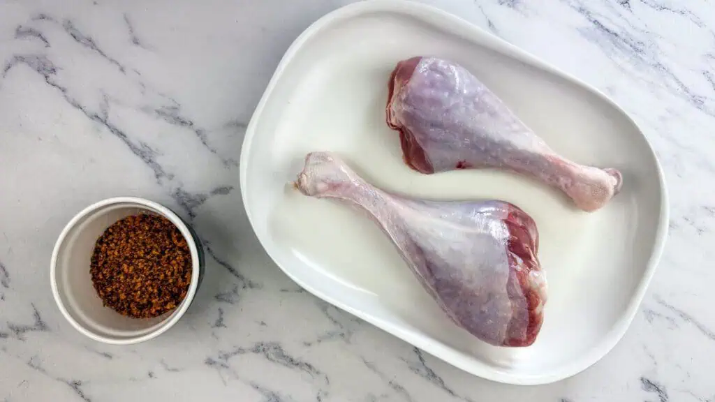 Two raw turkey legs on a white oval plate next to a small bowl of seasoning on a marble countertop.
