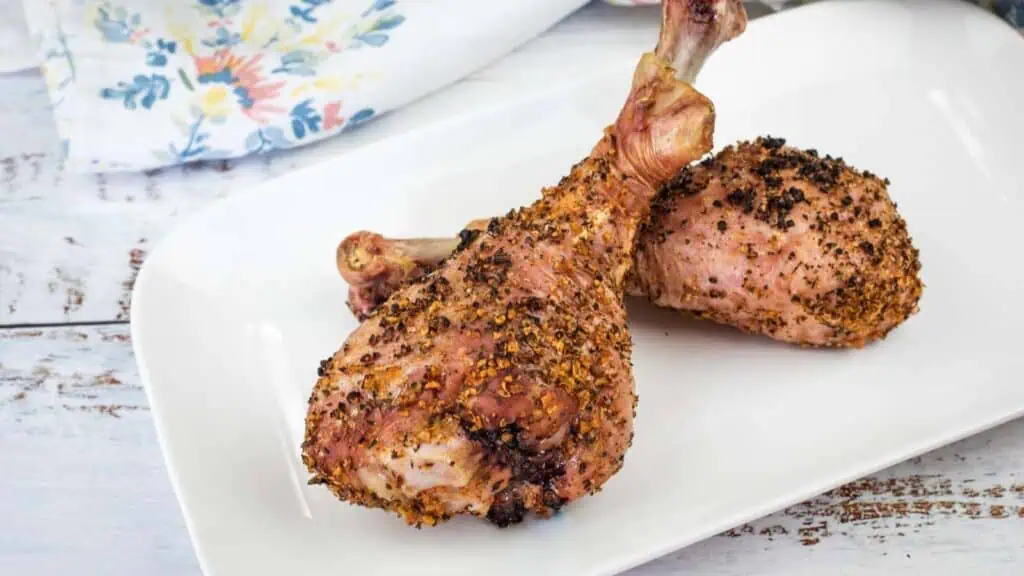 Two seasoned, baked turkey legs are arranged on a white rectangular plate atop a rustic wooden surface, with a floral napkin in the background.