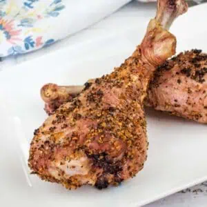 Two seasoned, baked turkey legs are arranged on a white rectangular plate atop a rustic wooden surface, with a floral napkin in the background.