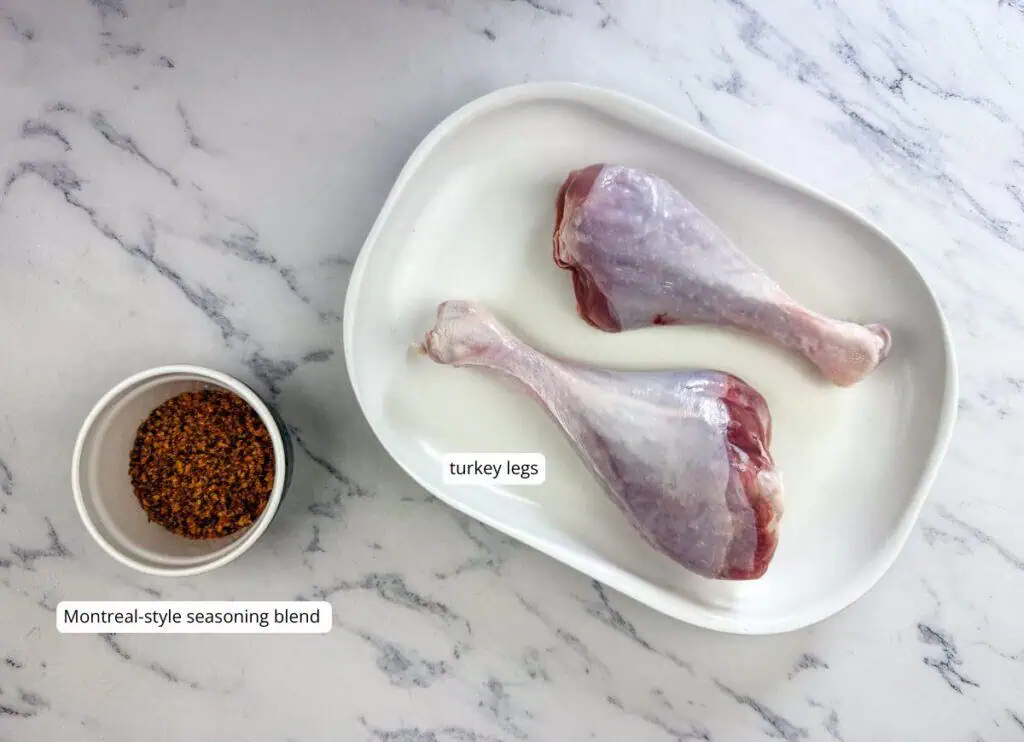 Two raw turkey legs on a white tray and a bowl of Montreal-style seasoning blend on a marble countertop. Labels identify the turkey legs and the seasoning.