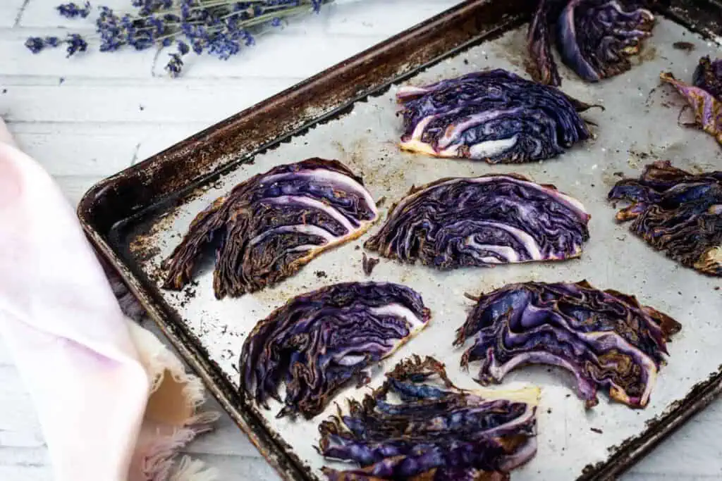 Roasted purple cabbage wedges arranged on a baking sheet, with some charred edges. Lavender and a light pink cloth are visible nearby.