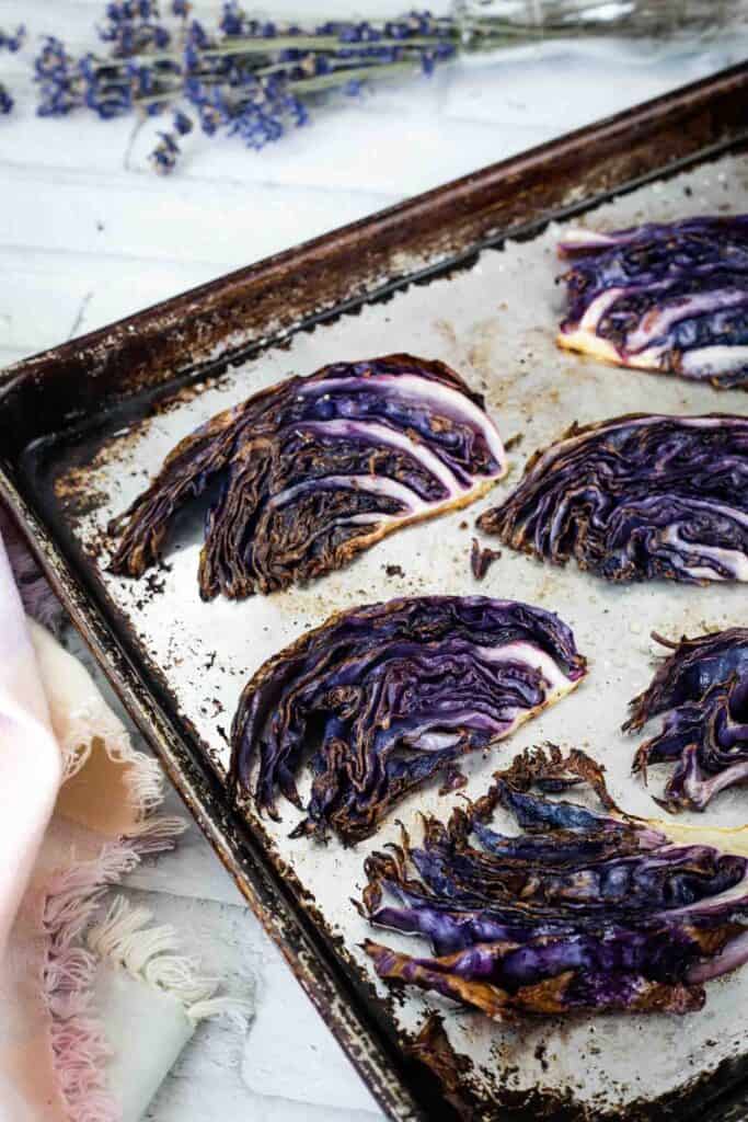 Roasted purple cabbage wedges on a metal baking sheet with a frayed cloth and lavender sprigs nearby.
