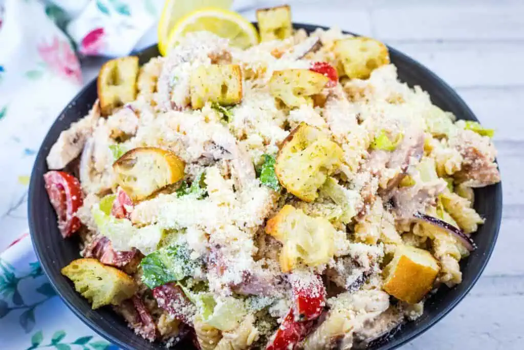 A bowl of chicken Caesar pasta salad topped with croutons, grated cheese, lettuce, cherry tomatoes, and lemon slices in the background.
