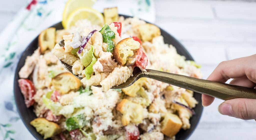 A hand holds a fork with pasta salad, croutons, lettuce, and tomatoes above a bowl of the same salad on a white surface.