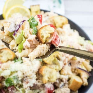 A hand holds a fork with pasta salad, croutons, lettuce, and tomatoes above a bowl of the same salad on a white surface.