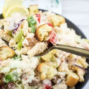 A hand holds a fork with pasta salad, croutons, lettuce, and tomatoes above a bowl of the same salad on a white surface.