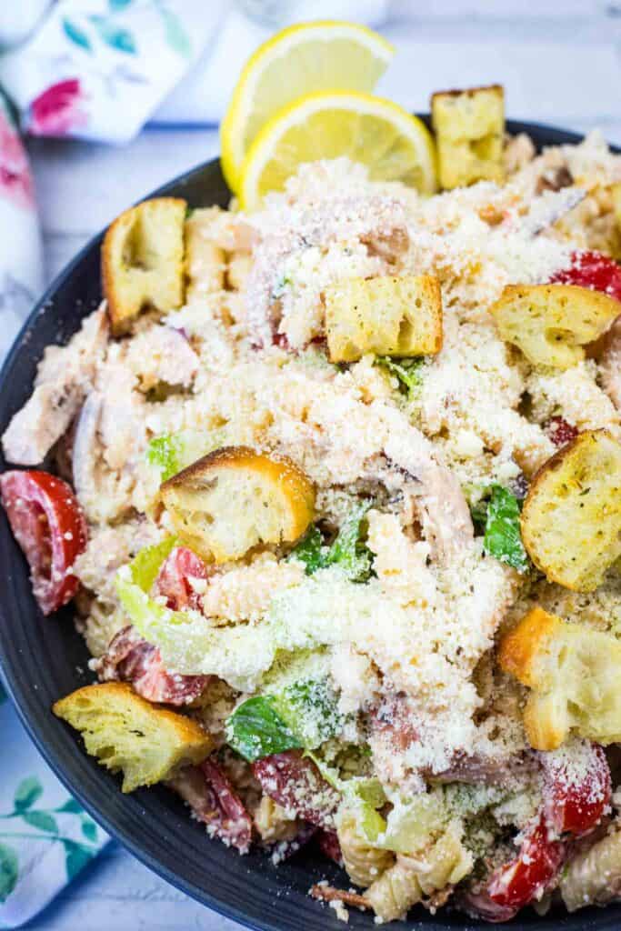 A bowl of chicken Caesar pasta salad topped with croutons, cherry tomatoes, lettuce, and grated parmesan cheese, garnished with lemon slices.