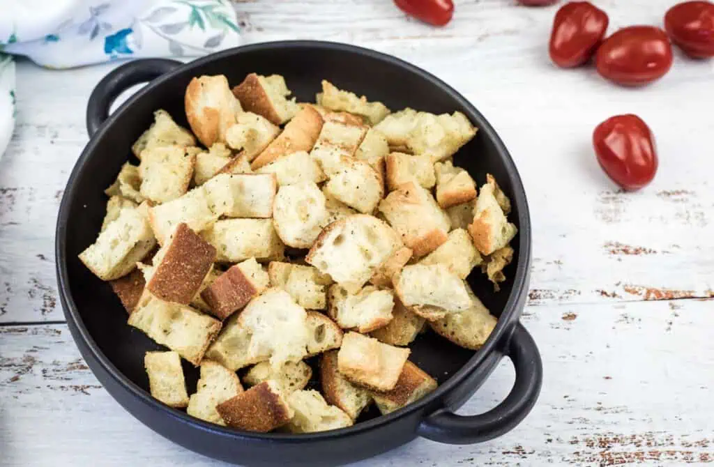 A black bowl filled with toasted bread croutons sits on a white wooden surface, with several cherry tomatoes scattered nearby.