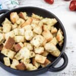 A black bowl filled with toasted bread croutons sits on a white wooden surface, with several cherry tomatoes scattered nearby.
