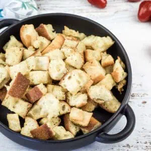 A black bowl filled with toasted bread croutons sits on a white wooden surface, with several cherry tomatoes scattered nearby.