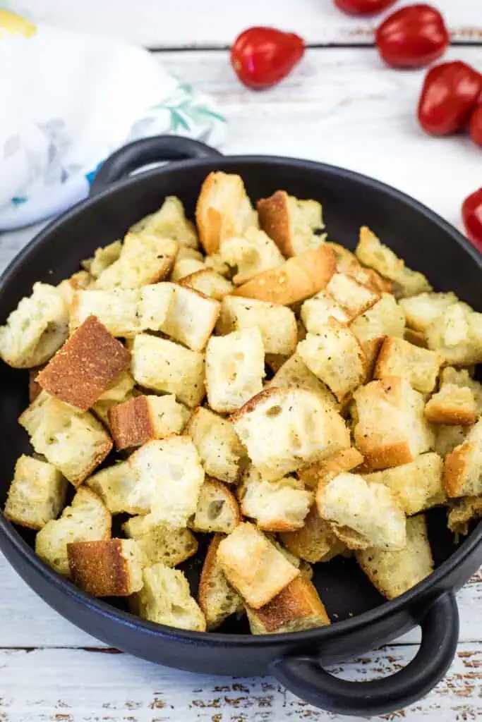 A black bowl filled with golden-brown croutons on a white wooden surface, with cherry tomatoes scattered nearby.