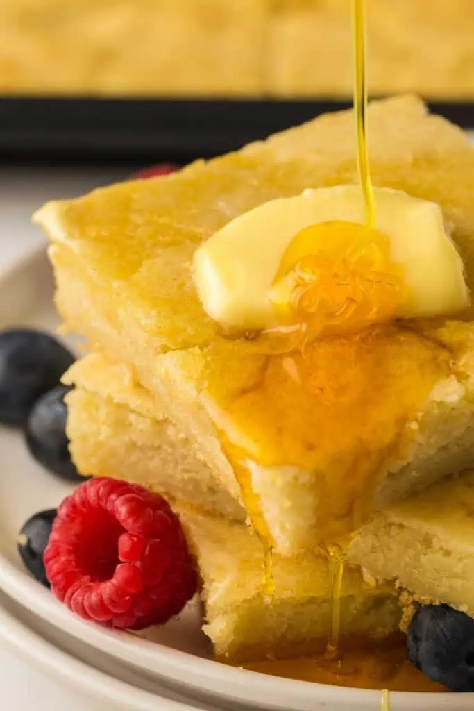 Two pancake squares topped with butter and syrup are stacked on a plate, garnished with fresh blueberries and a raspberry.