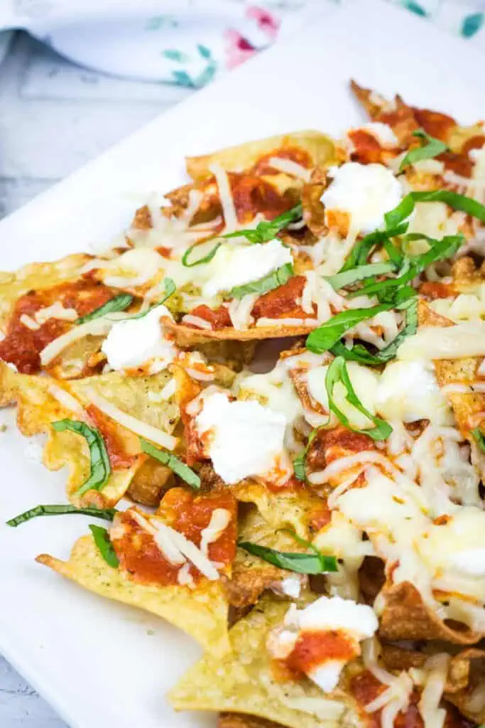 A plate of nachos topped with melted cheese, tomato sauce, ricotta, and sliced basil on a white dish.