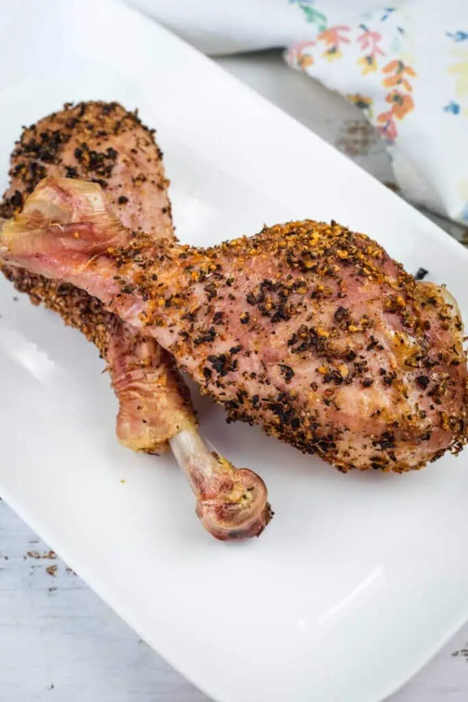 Two seasoned, cooked turkey legs are placed on a white rectangular plate, with a floral-patterned cloth partially visible in the background.