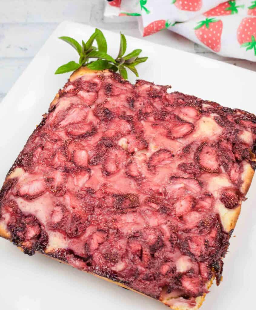 A square piece of strawberry-topped dessert sits on a white plate, garnished with a sprig of fresh mint in the corner.