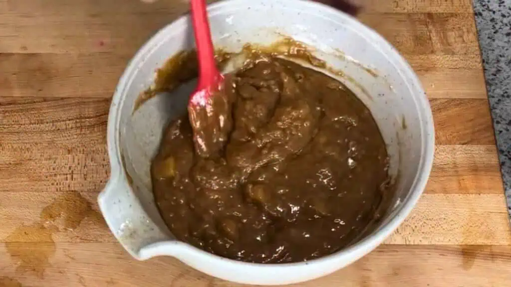 A white mixing bowl filled with thick brown batter is being stirred with a red spatula on a wooden surface.