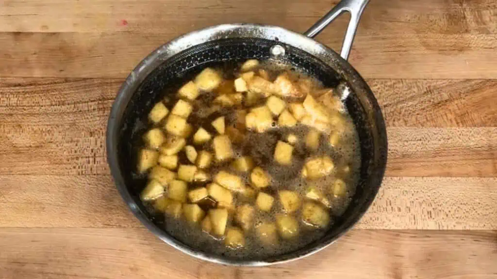 Diced apples simmering in a pan with brown liquid, likely sugar and spices, on a wooden surface.