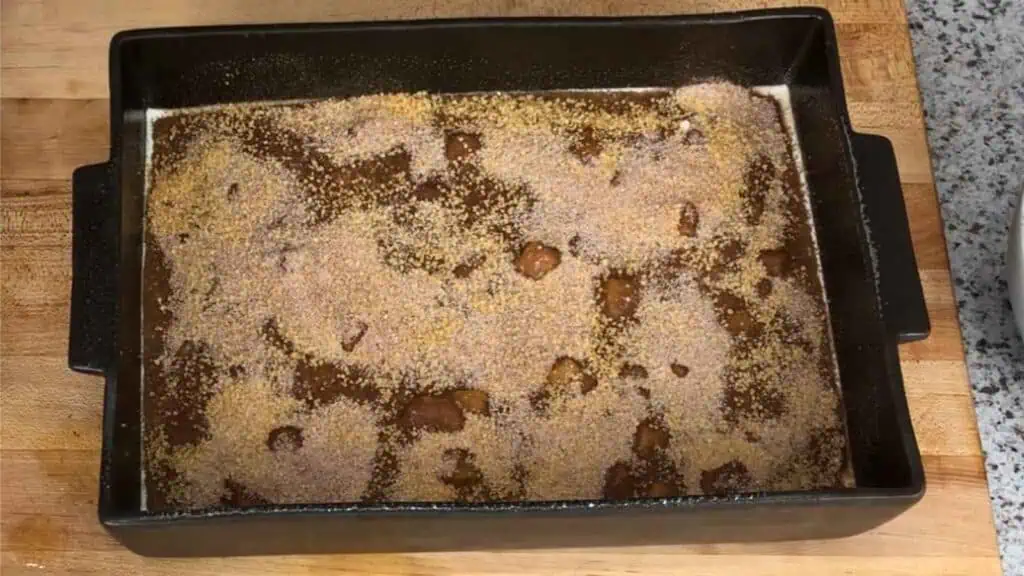 A rectangular baking dish filled with a brown mixture topped with a layer of granulated sugar, sitting on a wooden countertop.