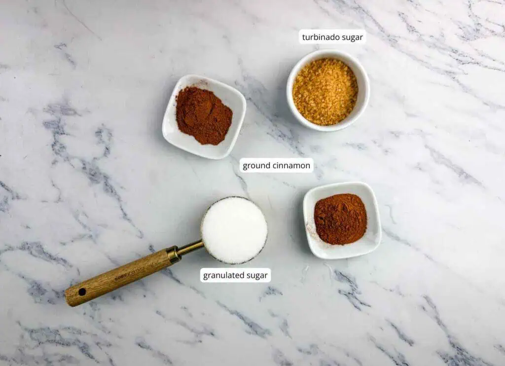 A measuring cup of granulated sugar, two bowls of ground cinnamon, and one bowl of turbinado sugar on a white marble surface.