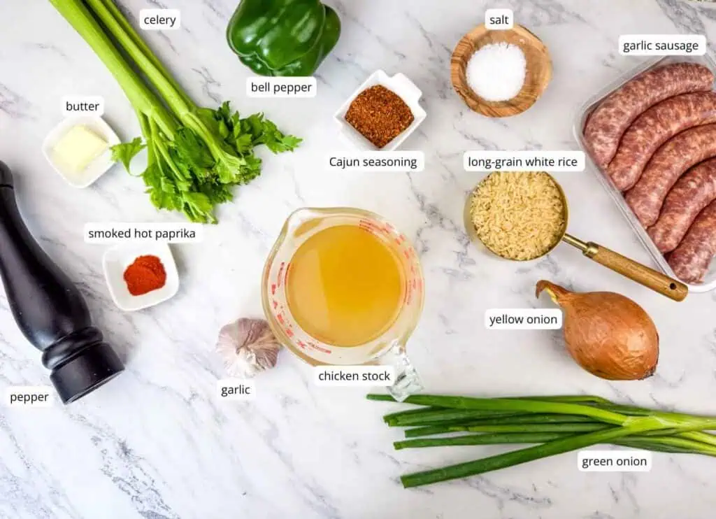 Labeled ingredients for cooking are arranged on a marble surface, including vegetables, spices, chicken stock, rice, butter, and sausage.