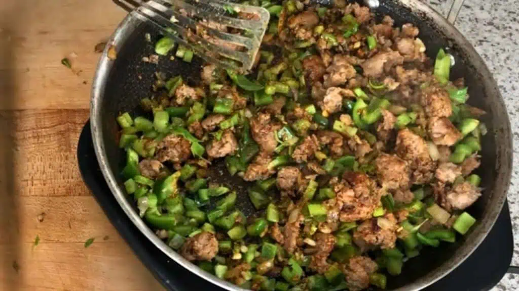 A frying pan on a stovetop contains chopped green bell peppers and browned ground meat being cooked together. A metal spatula rests in the pan.