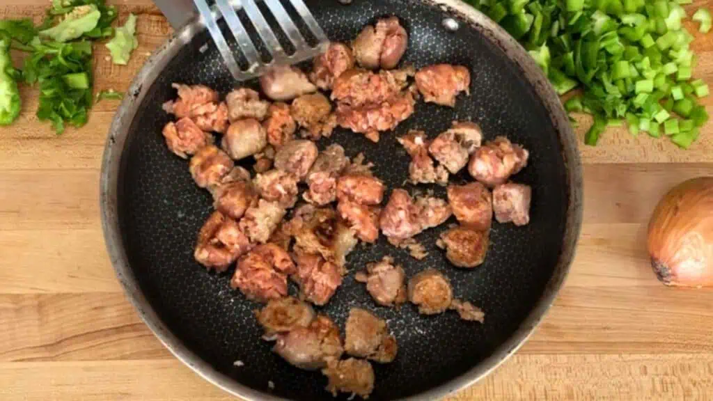 Small pieces of meat are being cooked in a frying pan, with chopped green vegetables and a whole onion on a wooden surface nearby.