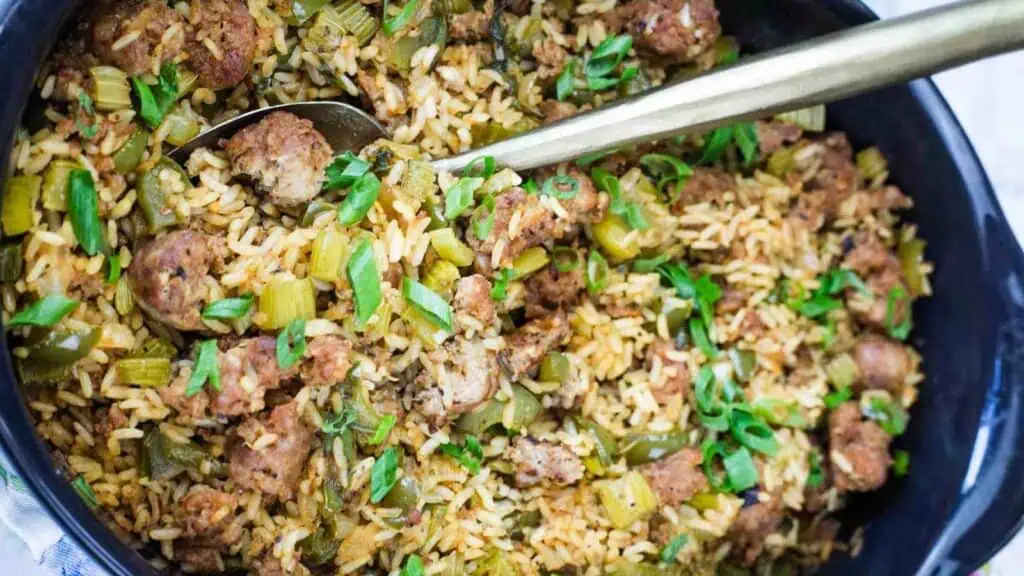 A skillet filled with cooked rice, sausage, chopped celery, green bell peppers, and green onions, with a spoon resting inside.