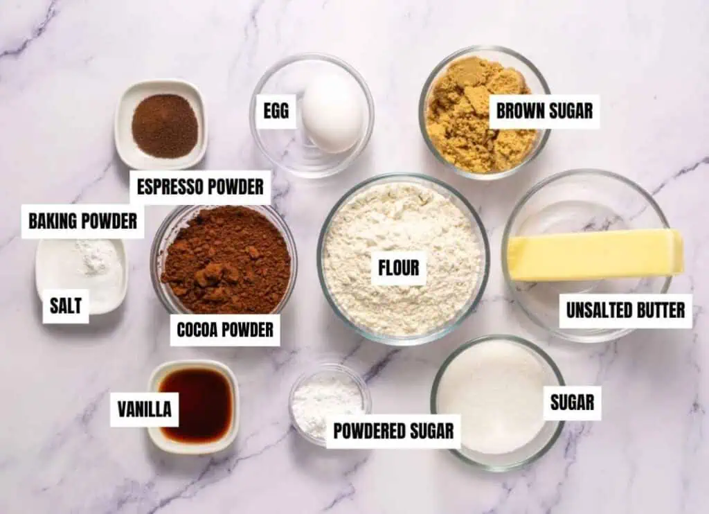 Various baking ingredients, including labeled bowls of flour, cocoa powder, sugars, butter, espresso powder, egg, vanilla, baking powder, and salt, are arranged on a white surface.