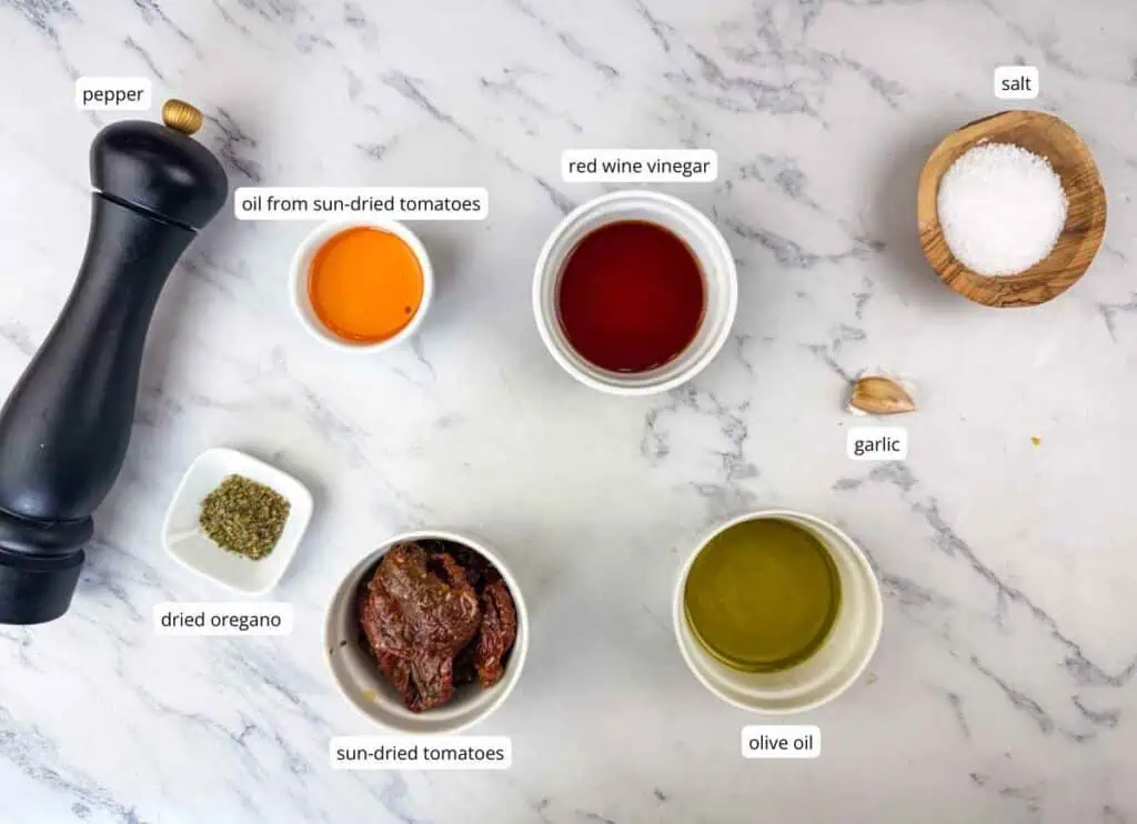 Ingredients for a recipe on a marble surface: black pepper grinder, sun-dried tomatoes, dried oregano, olive oil, red wine vinegar, salt, garlic, and oil from sun-dried tomatoes.