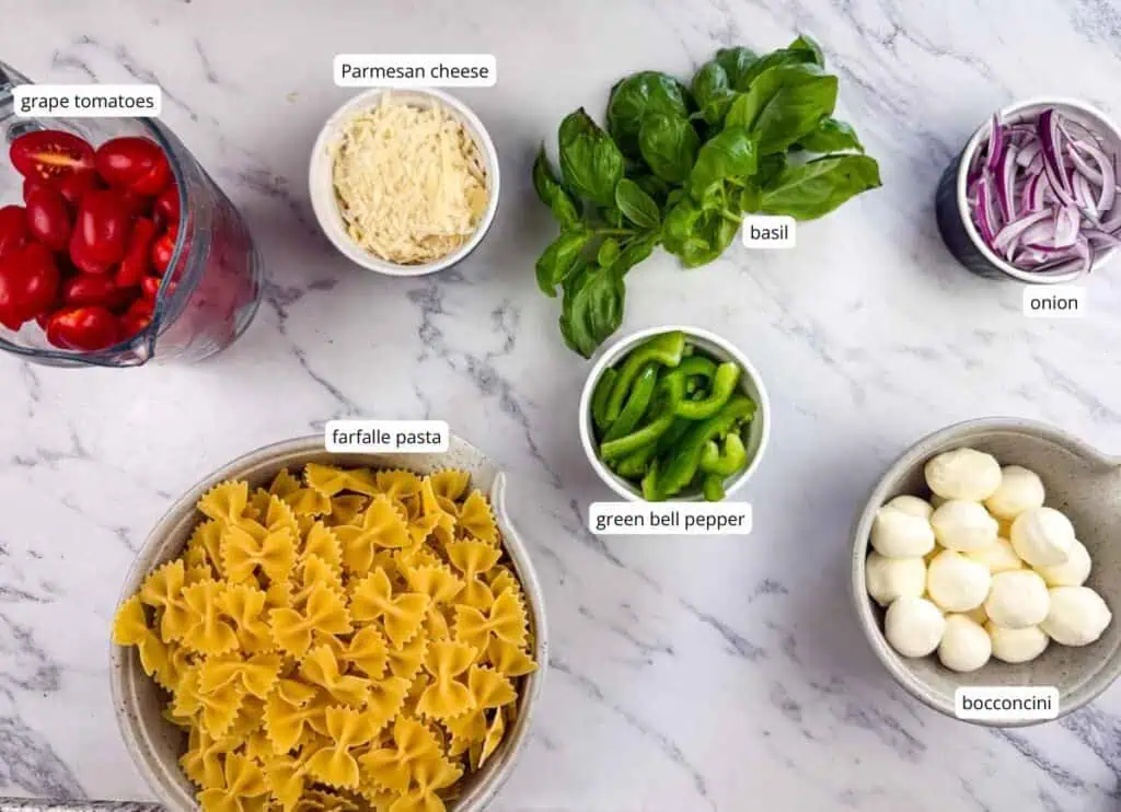 Bowls of grape tomatoes, Parmesan cheese, basil, onion, farfalle pasta, green bell pepper, and bocconcini are arranged on a marble surface.