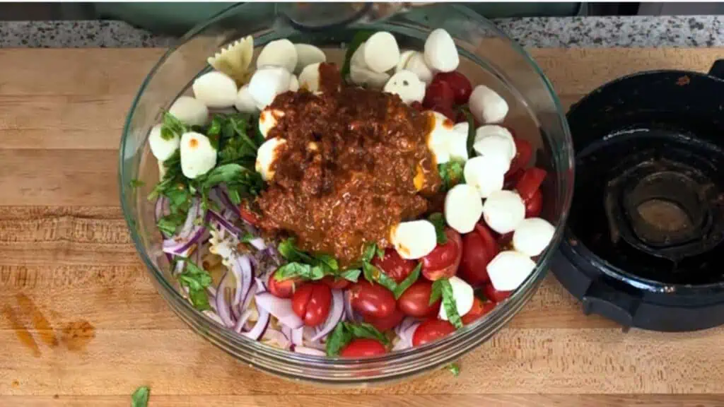Glass bowl with halved cherry tomatoes, mozzarella balls, red onion, basil, pasta, and a scoop of red sauce on a wooden cutting board next to a blender lid.
